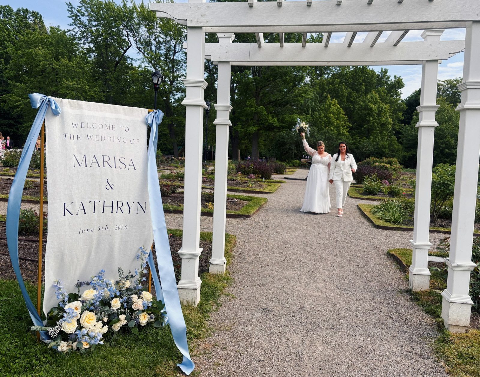 Brides walking hand in hand under a garden pergola, one holding her bouquet up in celebration
